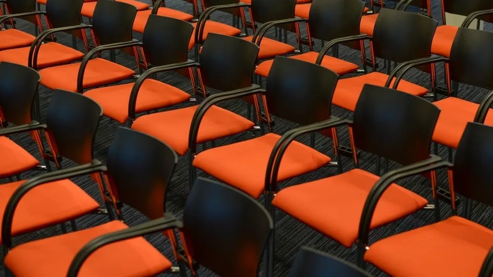 Des rangées de chaises orange et noires, vides, dans une salle de réunion.