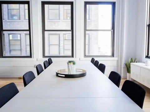 Une longue table blanche et ses chaises noires, devant de grandes fenêtres.